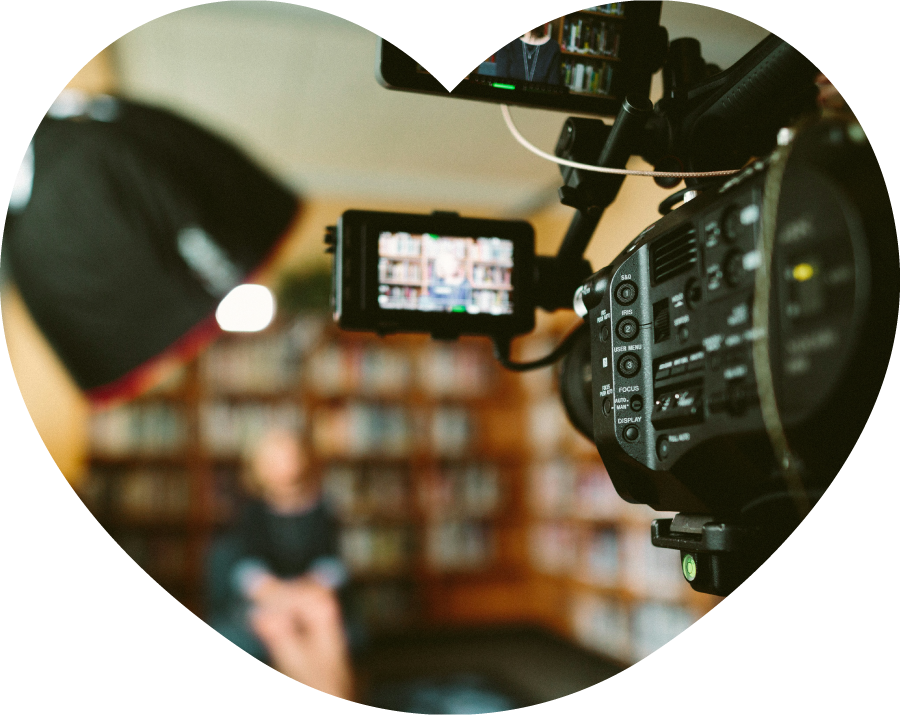 Man sieht im Vordergrund eine Videokamera und im Hintergrund verschwommen eine Frau, die vor einem Bücherregal sitzt.